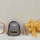 Gray backpack with a logo on a white surface next to a large yellow bow.