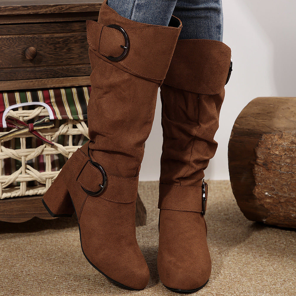 Brown knee-high boots with decorative buckles worn by a person, with a wooden drawer and basket in the background.