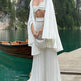 Woman in a white outfit standing on a dock by a lake with mountains in the background
