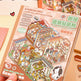 A hand holds a pink-covered sticker book titled 'STEREOSCOPIC LANDSCAPE CREATION STICKERS' with colorful illustrated collage scenes on the cover. The book appears to be the product being sold, photographed in a styled tabletop setting with small decorative props.