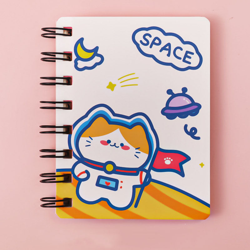 A spiral-bound notebook with a white cover featuring a cute cartoon cat in an astronaut space suit, surrounded by space-themed illustrations like a crescent moon, stars, a UFO, and a cloud that says 'SPACE'. The notebook is photographed on a solid pink background.