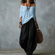 A woman wearing a light blue off-the-shoulder blouse and loose black harem pants, carrying a brown woven bag against a textured gray studio backdrop.
