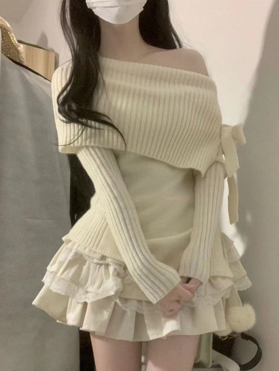 Woman wearing a beige off-shoulder knit cardigan over a dress with ruffled layers.