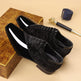 Black velvet shoes with floral patterns on a beige surface with a wooden box and pen in the background.