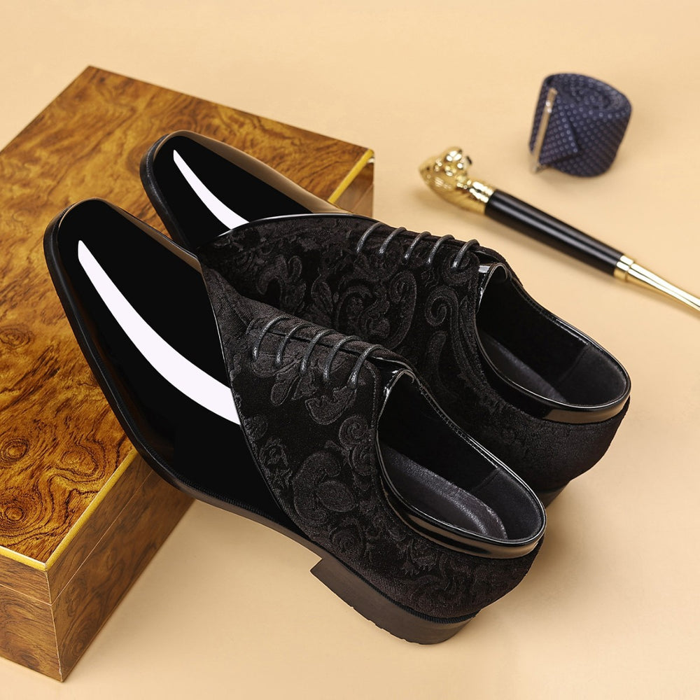 Black velvet shoes with floral patterns on a beige surface with a wooden box and pen in the background.