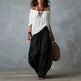 Woman wearing a white off-shoulder top and black wide-leg pants, holding a brown bag against a gray background.