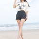A woman stands on a sandy beach wearing a light pink baseball cap, a cream graphic t-shirt that reads 'MORE SELF LOVE', black denim shorts, and pink and light blue athletic sneakers.