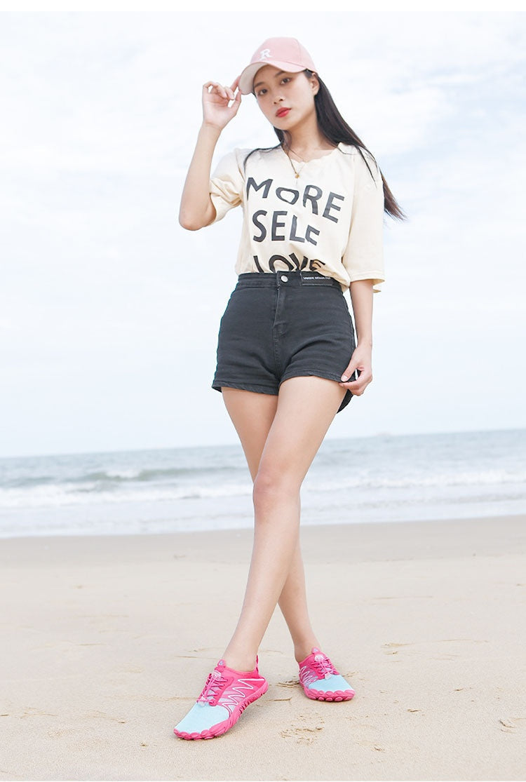 A woman stands on a sandy beach wearing a light pink baseball cap, a cream graphic t-shirt that reads 'MORE SELF LOVE', black denim shorts, and pink and light blue athletic sneakers.