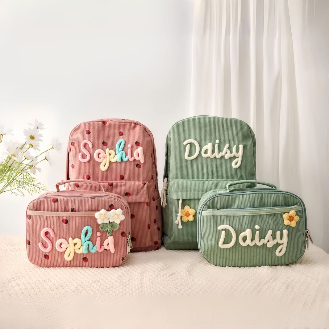 Four personalized corduroy backpacks and matching pouches in pink and green, decorated with embroidered names, floral accents, and small heart patterns, arranged on a white textured blanket with white curtains in the background.
