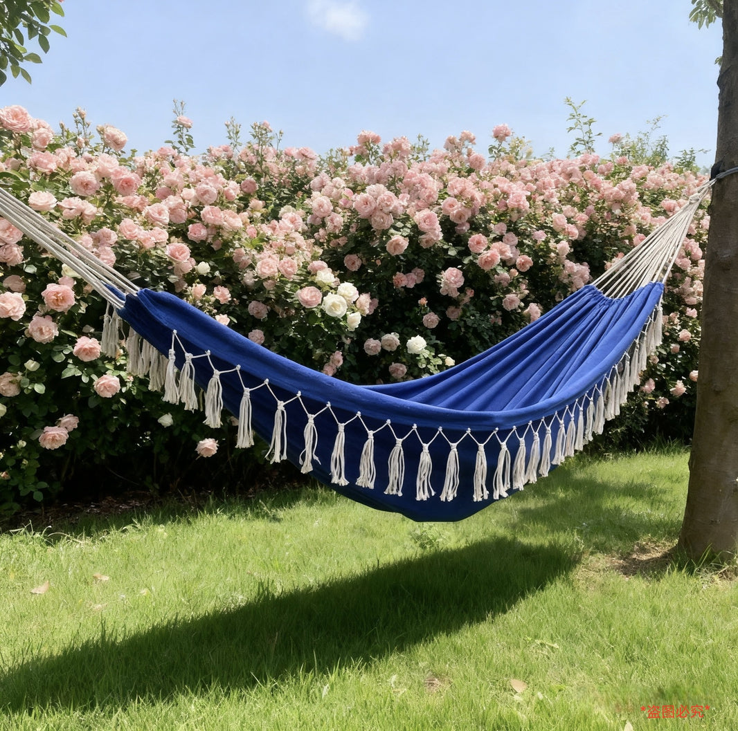 A blue hammock with white tassels is suspended between two trees in a garden setting, with green grass, a tree trunk, and a dense background of pink flowers under a blue sky.