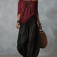 A woman models a bohemian-style outfit featuring a draped off-the-shoulder top in deep burgundy and wide-leg, voluminous black pants. She is accessorized with layered silver necklaces, bracelets, and large hoop earrings, and carries a woven brown tote bag against a textured gray studio backdrop.
