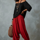 A woman modeling an off-the-shoulder black long-sleeve top and vibrant red baggy harem-style pants, accessorized with layered necklaces, bracelets, hoop earrings, and a woven straw bag, set against a textured gray studio backdrop.