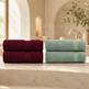Two stacks of folded bath towels displayed on a stone ledge in a softly lit bathroom setting. The left stack is a deep burgundy/wine color, and the right stack is a muted sage green.