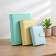 Three planner or notebook covers in mint green, pale yellow, and light blue are arranged on a tabletop in front of white window blinds. A cream pleated lamp is visible on the left, and a small jar candle and wooden coaster sit on the right.
