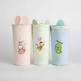 Three pastel-colored cylindrical cups with cat-ear lids are lined up on a light gray background. The left cup is light pink with a bunny and strawberry graphic, the middle is mint green with a tiger graphic, and the right is light blue with a green dinosaur graphic.