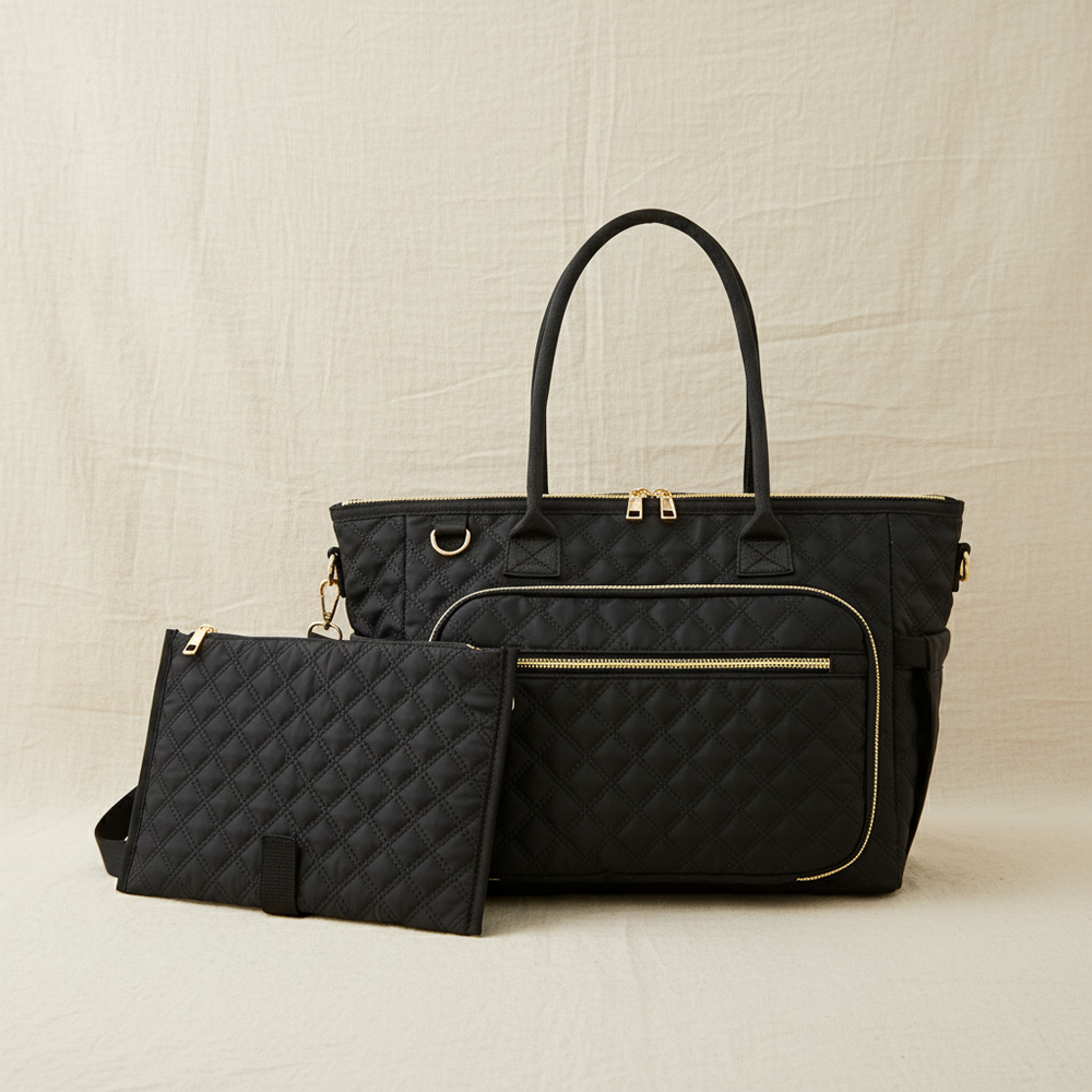 A black quilted diaper bag set with a matching zip pouch, featuring gold-tone hardware and dual top handles, displayed against a neutral beige fabric backdrop.