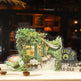 A decorative acrylic pop-up sign featuring a whimsical cafe scene with green trees, warm yellow window light, outdoor seating, a cup of coffee, and the phrase 'COFFEE IS ALWAYS A GOOD IDEA'. The sign is displayed on a tabletop with coffee beans against a blurred dark cafe interior background.