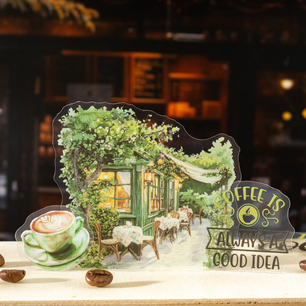 A decorative acrylic pop-up sign featuring a whimsical cafe scene with green trees, warm yellow window light, outdoor seating, a cup of coffee, and the phrase 'COFFEE IS ALWAYS A GOOD IDEA'. The sign is displayed on a tabletop with coffee beans against a blurred dark cafe interior background.